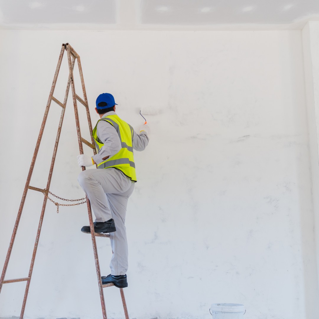 Professional Painter on Ladder Applying Paint to White Wall in Empty Room with Construction Safety Gear and Equipment